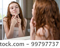 Teenage girl with red hair squeezes out pimples and acne on the face looking in the mirror in the bathroom of the house 99797737