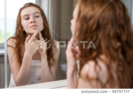 Teenage girl with red hair squeezes out pimples and acne on the face looking in the mirror in the bathroom of the house Teenage girl with red hair squeezes out pimples and acne on the face looking in the mirror in the bathroom of the house 99797737
