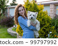 A teenage girl with red hair holds a pet, a white dog, outdoors in the summer 99797992