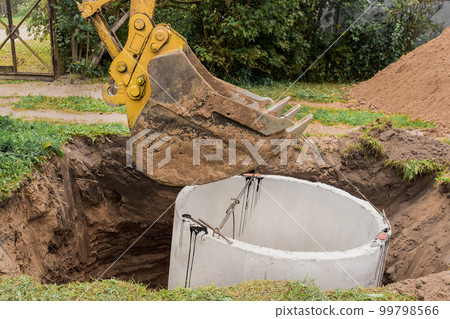 Excavator with a bucket, lowering into the pit on steel cables concrete sewer ring. Construction or repair of sewer home 99798566