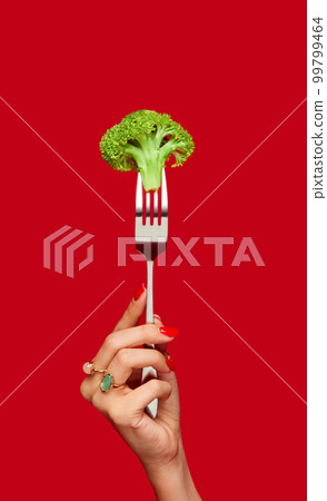 Female hand holding broccoli on fork against red studio background. Healthy eating. Food pop art photography. Female hand holding broccoli on fork against red studio background. Healthy eating. Food pop art photography. 99799464