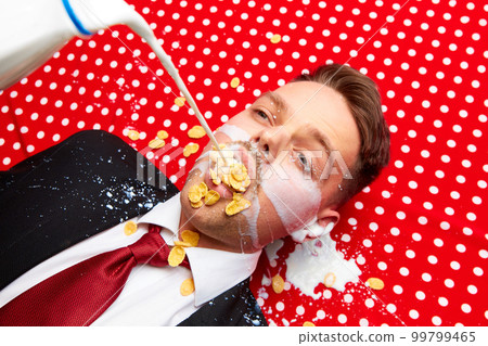 Young man lying on red vintage table cloth with corn flakes in mouth. Milk pouring inside mouth. Breakfast time. Food pop art photography. Young man lying on red vintage table cloth with corn flakes in mouth. Milk pouring inside mouth. Breakfast time. Food pop art photography. 99799465