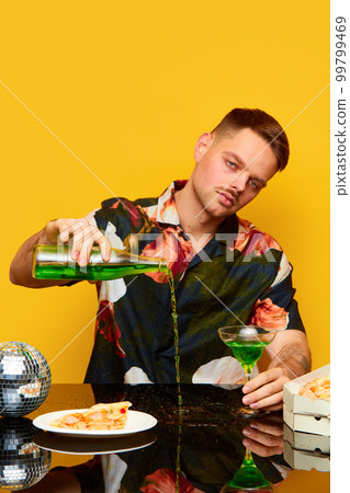 Young emotionless man in stylish shirt pouring cocktail on table against yellow studio background. Pizza slice and alcohol. Party. Food pop art 99799469
