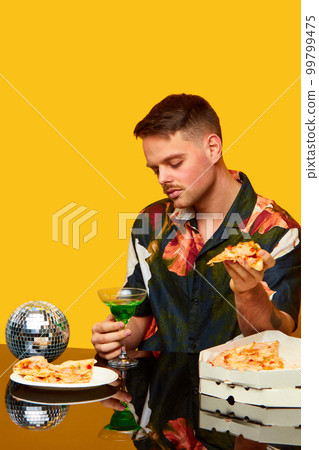 Food pop art photography. Young man in stylish shirt sitting at table, drinking cocktail and eating pizza against yellow studio background. Party 99799475