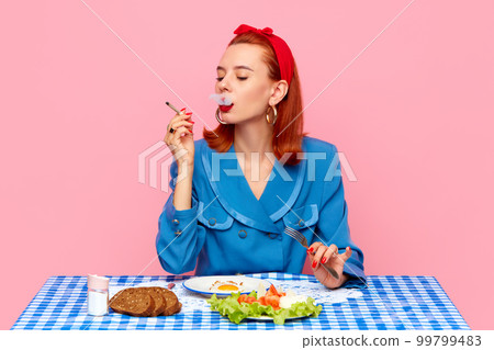 Stylish redhead woman sitting at table, smoking and having breakfast with fried eggs and salad against pink studio background. Food pop art photography. 99799483
