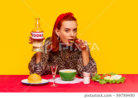 Beautiful stylish redhead woman sitting at table against yellow background and having dinner with cheese soup and drinking vodka. Food pop art photography 99799490