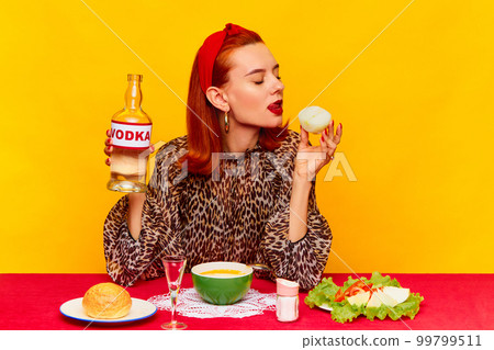 Food pop art photography. Beautiful redhead woman sitting at table against yellow background and having dinner, drinking vodka with onion. Food pop art photography. Beautiful redhead woman sitting at table against yellow background and having dinner, drinking vodka with onion. 99799511