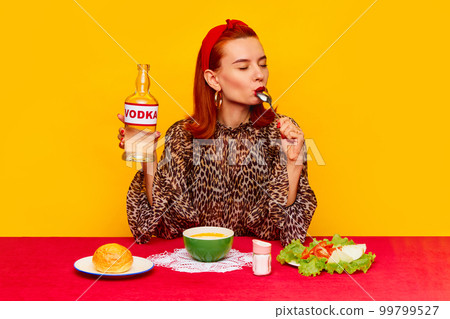 Stylish redhead woman sitting at table against yellow background and having dinner, eating soup, salad and drinking vodka. Food pop art photography. Stylish redhead woman sitting at table against yellow background and having dinner, eating soup, salad and drinking vodka. Food pop art photography. 99799527