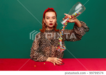Beautiful young redhead woman with serious face in stylish clothes pouring candies into wine glass over dark green studio background. 99799574