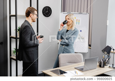 Couple of cheerful colleagues talking while standing at the flipchart in office with cups of coffee 99800605