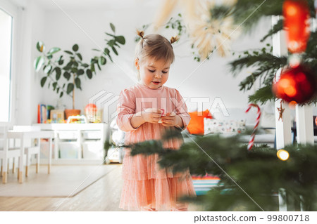 Beautiful caucasian baby gir with two little ponytails holding a Christmas ornament, looking at it with curiosity Beautiful caucasian baby gir with two little ponytails holding a Christmas ornament, looking at it with curiosity 99800718