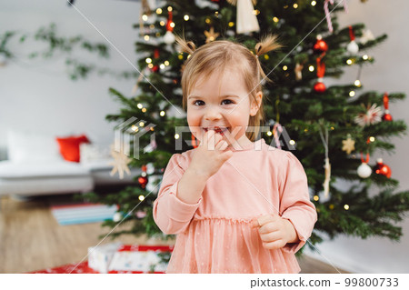 Cute little baby girl smiling at the camera chewing on her fingers, standing in front of the Christmas tree Cute little baby girl smiling at the camera chewing on her fingers, standing in front of the Christmas tree 99800733