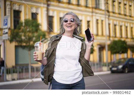 A Mature European Woman Enjoys Life Walking in The City. Biohacking Concept. Mature gray-haired modern european woman walks around the city in sunglasses, holding a mobile phone and a drink with a 99801462