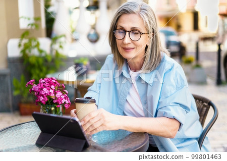 Silver-haired mature European woman sitting at an outdoor cafe, smiling with joy as she looks at her tablet PC. The image captures the happiness and contentment of a retiree enjoying leisure time and 99801463
