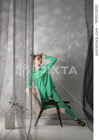 Stylish girl in green suit and black heels stands on gray wall background leaning on chair. Vertical frame Stylish girl in green suit and black heels stands on gray wall background leaning on chair. Vertical frame 99803800