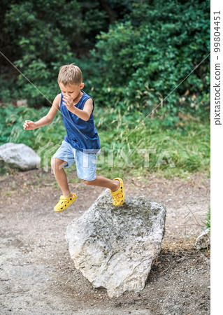 Active little boy jumps down from large rock in green summer park. Cute preschooler kid in casual clothes plays in wild nature park during walk Active little boy jumps down from large rock in green summer park. Cute preschooler kid in casual clothes plays in wild nature park during walk 99804451