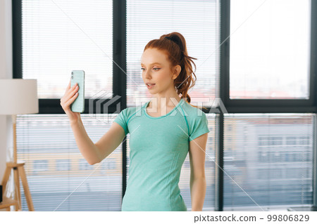 Portrait of sporty female vlogger in activewear recording live tutorial video to followers in light room on background of window. Young pretty woman making video call via mobile phone at home. 99806829
