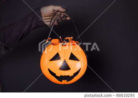 A bucket of candy in the form of a pumpkin lantern Jack on a black background in a hand in a mesh glove. Accessories for Halloween. A bucket of candy in the form of a pumpkin lantern Jack on a black background in a hand in a mesh glove. Accessories for Halloween. 99810295