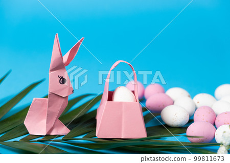 An Easter bunny figurine made of paper, origami, and a basket with eggs on a blue background, place for text 99811673