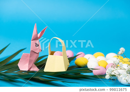 An Easter bunny figurine made of paper, origami, and a basket with eggs on a blue background, place for text 99811675