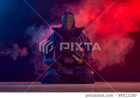 Man, professional kendo athlete, combat sportsman posing with shinai sword against gradient dark background in neon light with smoke Man, professional kendo athlete, combat sportsman posing with shinai sword against gradient dark background in neon light with smoke 99812280