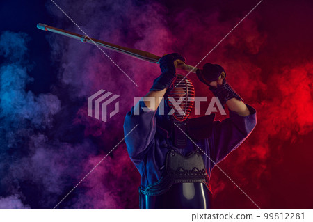 Man, professional kendo athlete, combat sportsman posing with shinai sword against gradient dark background in neon light with smoke Man, professional kendo athlete, combat sportsman posing with shinai sword against gradient dark background in neon light with smoke 99812281