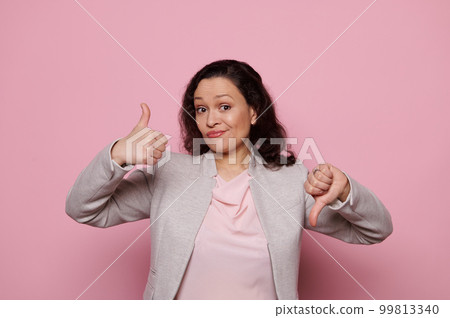 Doubtful multiracial brunette woman in elegant gray jacket, gestures with her hands, analyzes the pros and cons, shows thumbs up and down, thoughtfully looking at camera, isolated on pink background Doubtful multiracial brunette woman in elegant gray jacket, gestures with her hands, analyzes the pros and cons, shows thumbs up and down, thoughtfully looking at camera, isolated on pink background 99813340
