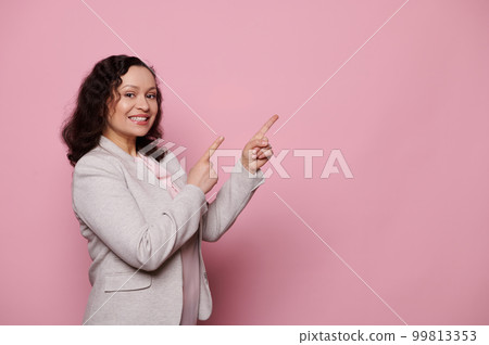 Beautiful middle-aged ethnic woman, business person, manager in light gray formal jacket, points forefingers at copy space for ads on pink isolated background, smiling a toothy smile looking at camera Beautiful middle-aged ethnic woman, business person, manager in light gray formal jacket, points forefingers at copy space for ads on pink isolated background, smiling a toothy smile looking at camera 99813353
