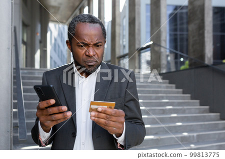 Problems with online shopping, ordering. A worried senior African American man in a suit stands outside an office center. He is holding a credit card and a phone. Problems with online shopping, ordering. A worried senior African American man in a suit stands outside an office center. He is holding a credit card and a phone. 99813775