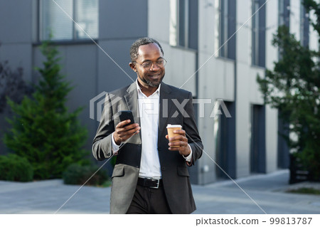 Coffee break. A successful African American businessman in a business suit is standing on the street near a skyscraper in a suit and glasses, holding a cup of coffee to go and using a mobile phone. 99813787