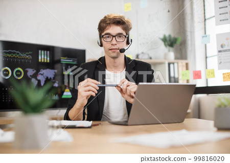 Young businessman in headset using laptop computer during video call working in office. 99816020