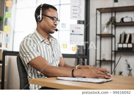 Busy african man using headset in striped shirt sitting at office and using wireless laptop. 99816092