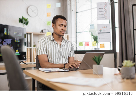 Busy african american man in striped shirt sitting at office and using wireless laptop. 99816113