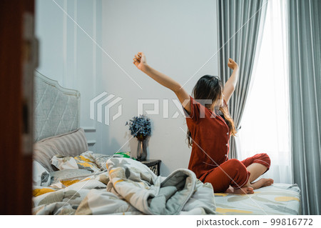 long haired asian young woman sitting in bed stretching 99816772