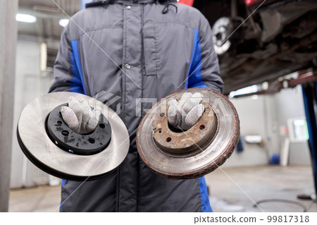 The mechanic holds old rusty brake disc and new disc. Change the old to new brake disc on car in a garage. Auto repair concept. The mechanic holds old rusty brake disc and new disc. Change the old to new brake disc on car in a garage. Auto repair concept. 99817318