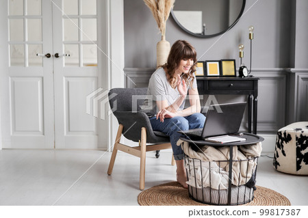 Woman waving hand has video call laptop computer online sitting in armchair at home. Hello there. Pleasant young female teacher or tutor sitting at home in cozy room before laptop  99817387