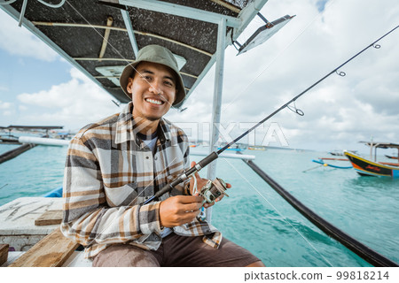 Asian angler smiles while holding a fishing rod on boat 99818214