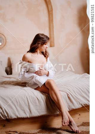 an attractive slender naked woman in a white shirt sit on the bed. 99821581