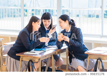 A high school girl talking to her classmate while looking at her smartphone in the classroom A high school girl talking to her classmate while looking at her smartphone in the classroom 99835176