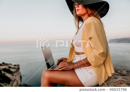 Digital nomad, Business woman working on laptop by the sea. Pretty lady typing on computer by the sea at sunset, makes a business transaction online from a distance. Freelance, remote work on vacation 99835796