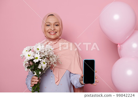 Smiling Muslim woman in pink hijab, holds bunch of wildflowers, shows smartphone with blank screen, with free ad space for your mobile application, isolated background with pink pastel helium balloons Smiling Muslim woman in pink hijab, holds bunch of wildflowers, shows smartphone with blank screen, with free ad space for your mobile application, isolated background with pink pastel helium balloons 99842224