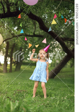 A funny birthday girl in a blue dress and a pink festive cap is standing under a holiday garland A funny birthday girl in a blue dress and a pink festive cap is standing under a holiday garland 99851289