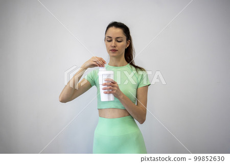 A young, slim woman in sportswear twisting the lid of a sports nutrition shaker against a gray background. Copy space. 99852630