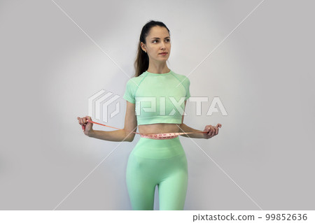Young slender woman in sportswear measuring her waist with a tape measure against a gray background. Copy Space. The concept of diet and weight loss. Young slender woman in sportswear measuring her waist with a tape measure against a gray background. Copy Space. The concept of diet and weight loss. 99852636