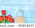 Red tulips and a paper craft bag on a blue wooden background, top view. 99854025