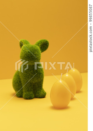 Green Easter Bunny with three yellow candles of eggs shape on matching monochrome orange background. Minimal Happy Easter holiday concept. Creative festive Easter greeting card, copy space, vertical. Green Easter Bunny with three yellow candles of eggs shape on matching monochrome orange background. Minimal Happy Easter holiday concept. Creative festive Easter greeting card, copy space, vertical. 99855087