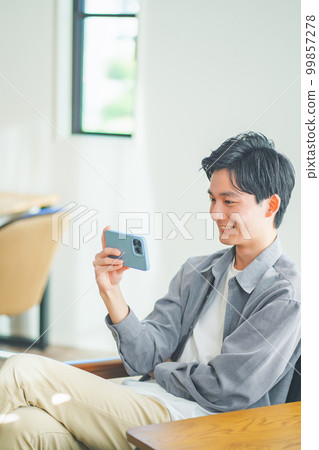 Young man sitting on sofa and watching videos on smartphone Young man sitting on sofa and watching videos on smartphone 99857278