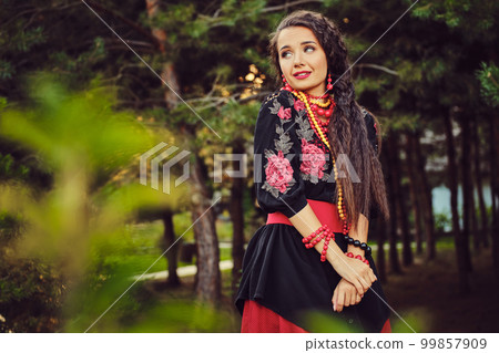 Brunette girl in a white ukrainian authentic national costume and a wreath of flowers is posing in a wood. Close-up. 99857909