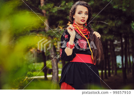 Brunette girl in a white ukrainian authentic national costume and a wreath of flowers is posing in a wood. Close-up. 99857910