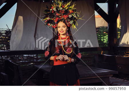 Brunette girl in a white ukrainian authentic national costume and a wreath of flowers is posing against a terrace. Brunette girl in a white ukrainian authentic national costume and a wreath of flowers is posing against a terrace. 99857950